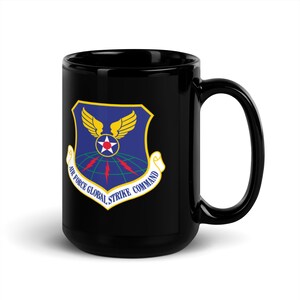 Peut inclure: Mug en céramique noire avec un contour blanc d'un bouclier bleu avec une étoile jaune et des ailes. Le texte "Air Force Global Strike Command" est écrit autour du bouclier.