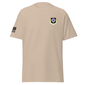 Könnte beinhalten: Ein beiges T-Shirt mit einer schwarz-weißen amerikanischen Flagge am linken Ärmel und einem schwarz-gelben Aufnäher auf der rechten Brust. Der Aufnäher zeigt einen Stern, Flügel und den Text "341st Space Wing".