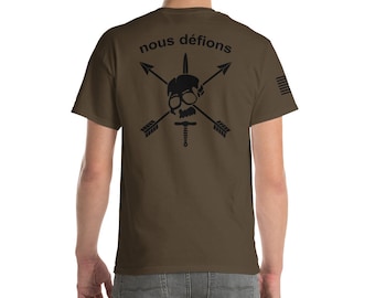 Puede incluir: Camiseta verde oliva con un gráfico negro en la espalda que presenta una calavera, flechas y una espada. El texto "nous défions" está encima del gráfico. Una bandera estadounidense está en la manga derecha.