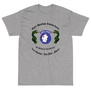 Puede incluir: Una camiseta gris con un gráfico de un logotipo de un centro de entrenamiento de operaciones en la jungla. El logotipo presenta dos serpientes verdes que flanquean un círculo azul con un barco blanco y una cruz roja. El texto "Jungle Expert" está escrito en una pancarta negra sobre el círculo. El texto "No obstacle too difficult" está escrito debajo del círculo. El texto "Fort Sherman - Toro Point - Panama" está escrito debajo del texto.