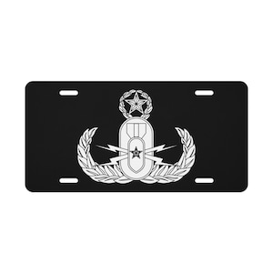 May include: Black license plate with a white emblem depicting a star, lightning bolts, and a wreath. The emblem is centrally positioned, with four mounting holes. The design is a military-themed graphic.