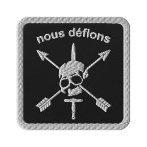 May include: Black and white embroidered patch with a skull, crossed arrows, and a sword. The text "nous défions" is above the skull.