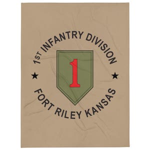 May include: Tan blanket featuring the text "1st Infantry Division Fort Riley Kansas" in black, encircling a green and red emblem. Two black stars flank the emblem. The blanket is rectangular.
