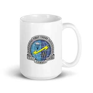 Może przedstawiać: Biały kubek ceramiczny z czarno-białym nadrukiem wojskowego emblematu. Emblemat przedstawia glob z błyskawicą, spadochronem i tekstem "First There U.S.A.F. Combat Control".