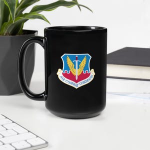 May include: Black ceramic mug featuring the Air Combat Command emblem. The emblem is a shield with a sword, wings, and text that reads "Air Combat Command". The mug is on a white surface with a plant and a keyboard in the background.