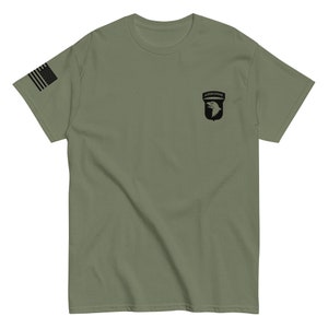 May include: A green t-shirt with a black and white 101st Airborne Division patch on the right chest. The shirt has a small American flag on the left sleeve.