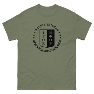 Peut inclure: Un t-shirt vert avec un graphique circulaire noir. Le graphique représente un bouclier avec le texte "IFOR" sur le côté gauche et "IFOR" sur le côté droit. Le cercle comprend également le texte "Bosnia Veteran", "Operation Joint Endeavor", et cinq étoiles.