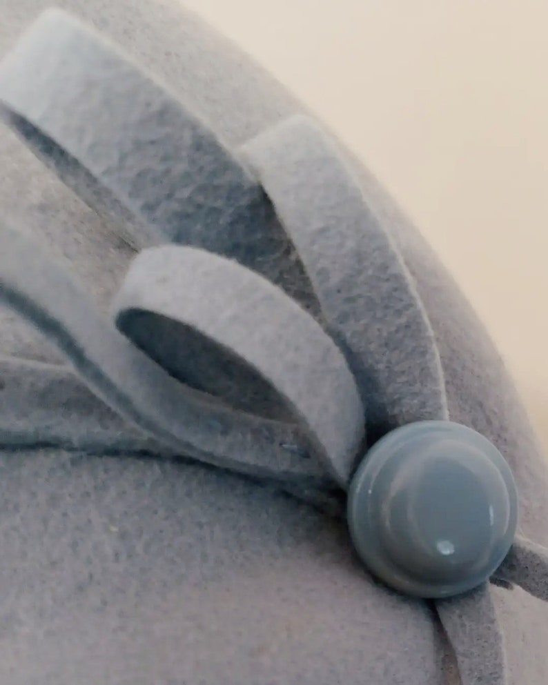 Pu&ograve; includere: Primo piano di un cappello in feltro azzurro con un fiocco decorativo e un bottone abbinato. La consistenza morbida e il design semplice del cappello creano un look classico ed elegante. Il bottone ha un diametro di circa 2,5 cm.