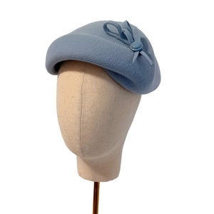 Pu&ograve; includere: Un basco azzurro con un piccolo fiocco e un dettaglio a bottone. Il cappello &egrave; esposto su una testa di manichino su sfondo bianco. La consistenza morbida e il design classico del cappello suggeriscono uno stile vintage o retr&ograve;.