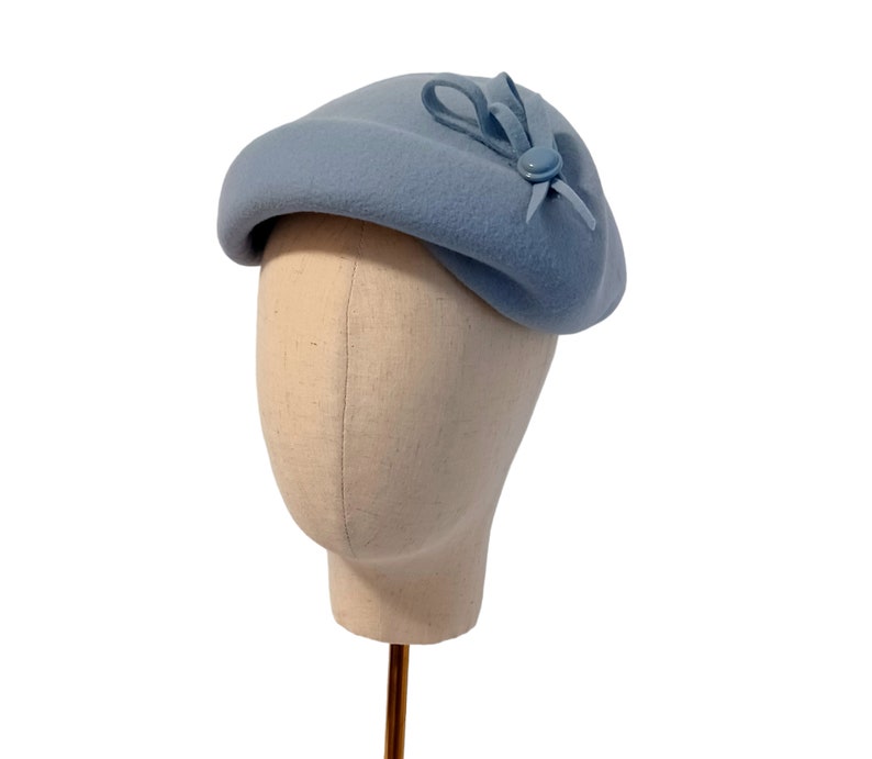 Pu&ograve; includere: Un basco azzurro con un piccolo fiocco e un dettaglio a bottone. Il cappello &egrave; esposto su una testa di manichino su sfondo bianco. La consistenza morbida e il design classico del cappello suggeriscono uno stile vintage o retr&ograve;.