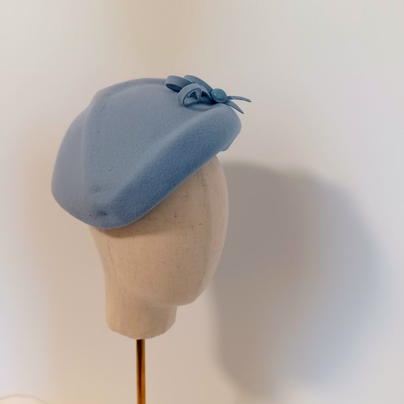 Pu&ograve; includere: Un cappello in stile basco azzurro con un piccolo fiocco e un dettaglio decorativo. Il cappello &egrave; esposto su una testa di manichino. Il cappello &egrave; realizzato in un materiale morbido ed &egrave; di colore pieno.
