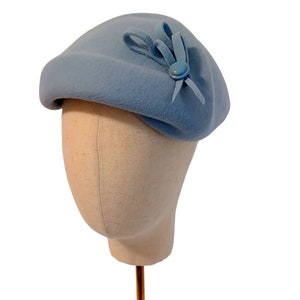 Pu&ograve; includere: Un cappello a basco blu chiaro con un piccolo fiocco e un bottone. Il cappello &egrave; realizzato in un materiale morbido ed &egrave; esposto su una testa di manichino. Un accessorio classico.