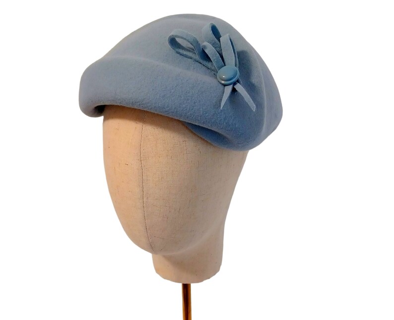 Pu&ograve; includere: Un cappello a basco blu chiaro con un piccolo fiocco e un bottone. Il cappello &egrave; realizzato in un materiale morbido ed &egrave; esposto su una testa di manichino. Un accessorio classico.