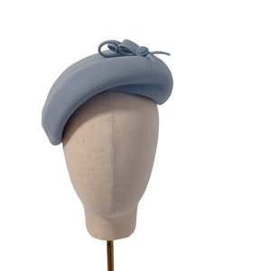 Pu&ograve; includere: Basco azzurro chiaro con un piccolo fiocco e dettaglio a bottone. Il cappello &egrave; esposto su una testa di manichino su sfondo bianco. Il basco &egrave; di colore pieno e ha una forma classica.