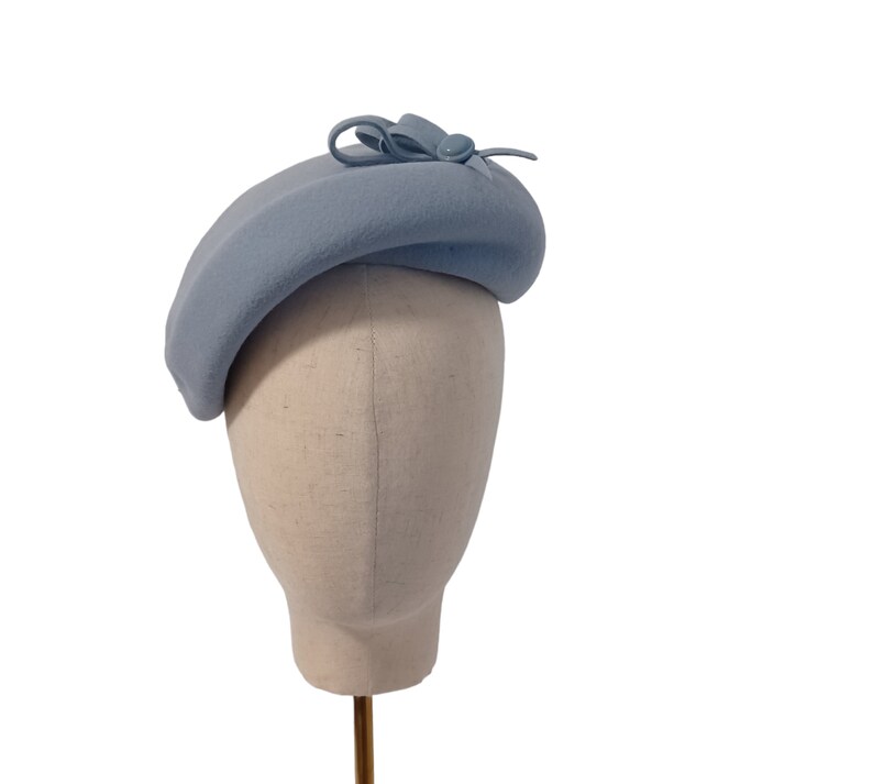 Pu&ograve; includere: Basco azzurro chiaro con un piccolo fiocco e dettaglio a bottone. Il cappello &egrave; esposto su una testa di manichino su sfondo bianco. Il basco &egrave; di colore pieno e ha una forma classica.