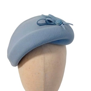 Pu&ograve; includere: Un cappello a forma di basco azzurro chiaro con un piccolo fiocco e un bottone. Il cappello &egrave; realizzato in un materiale morbido ed &egrave; esposto su una testa di manichino su sfondo bianco. Un accessorio classico.