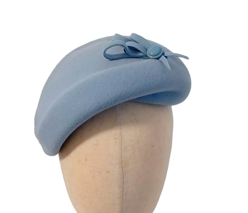 Pu&ograve; includere: Un cappello a forma di basco azzurro chiaro con un piccolo fiocco e un bottone. Il cappello &egrave; realizzato in un materiale morbido ed &egrave; esposto su una testa di manichino su sfondo bianco. Un accessorio classico.