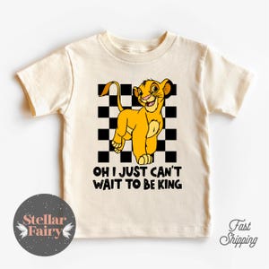 May include: Cream-colored toddler t-shirt featuring a cartoon lion cub with the text "OH I JUST CAN'T WAIT TO BE KING." The lion is yellow with a black and white checkered background. The shirt has short sleeves.