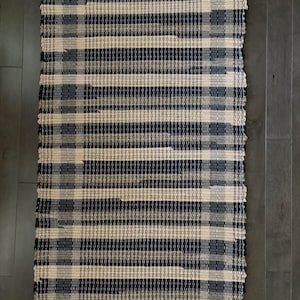 May include: A rectangular woven rug with alternating horizontal stripes of navy blue and beige. The rug has a textured appearance and a fringed edge. The rug is made of a durable material and is suitable for high-traffic areas.