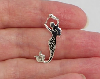May include: A silver mermaid charm with a black enamel design. The mermaid is holding her tail in one hand and has long flowing hair.