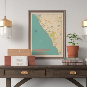 Può includere: Una mappa vintage incorniciata della costa della contea di Orange in California, con una scatola di legno e una pianta in vaso su un tavolo di legno marrone scuro con due cassetti. La mappa è in una cornice marrone chiaro con uno sfondo blu chiaro e testo rosso e giallo.