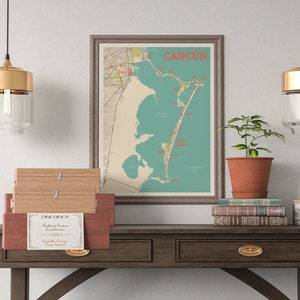 May include: A vintage map of Cancun, Mexico, framed in a brown frame, hangs on a white wall above a wooden table with two drawers. Two gold pendant lights with clear glass shades hang above the table. A potted plant sits on the table next to a stack of books and a small candle.