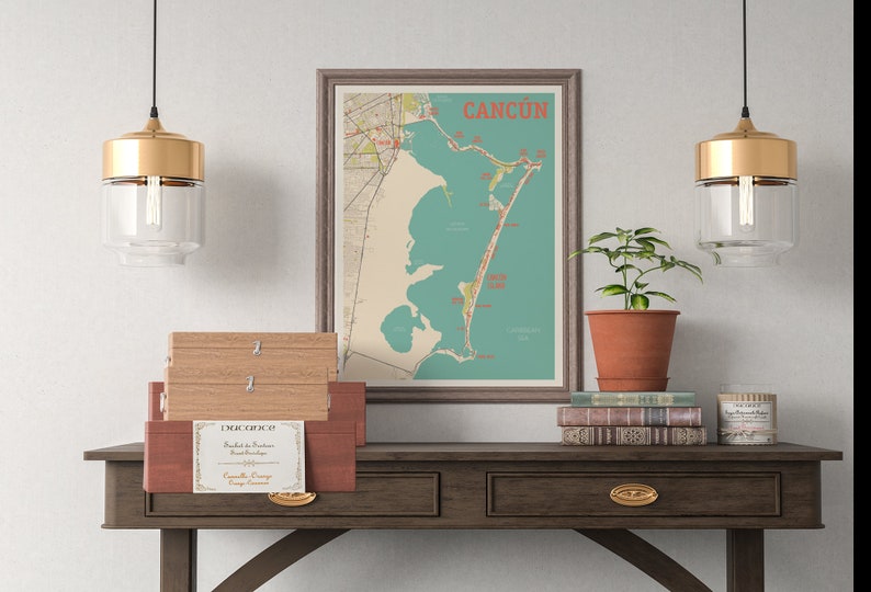 May include: A vintage map of Cancun, Mexico, framed in a brown frame, hangs on a white wall above a wooden table with two drawers. Two gold pendant lights with clear glass shades hang above the table. A potted plant sits on the table next to a stack of books and a small candle.