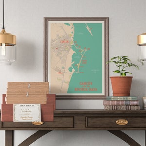 May include: A framed map of Cancun and the Riviera Maya with red and teal accents. The map is hanging on a white wall above a dark brown wooden table with two drawers. There are three wooden boxes stacked on the table, and a potted plant and a stack of books are on the table to the right of the map. Two gold pendant lights with clear glass shades hang from the ceiling above the table.