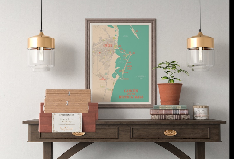 May include: A framed map of Cancun and the Riviera Maya with red and teal accents. The map is hanging on a white wall above a dark brown wooden table with two drawers. There are three wooden boxes stacked on the table, and a potted plant and a stack of books are on the table to the right of the map. Two gold pendant lights with clear glass shades hang from the ceiling above the table.