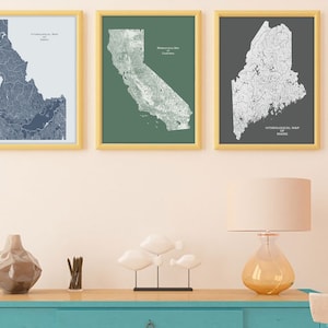 May include: Three framed maps of Idaho, California, and Maine. Each map is a black and white line drawing of the state's hydrological features, printed on a colored background. The Idaho map is on a light blue background, the California map is on a green background, and the Maine map is on a dark gray background. The maps are hanging on a light peach wall.