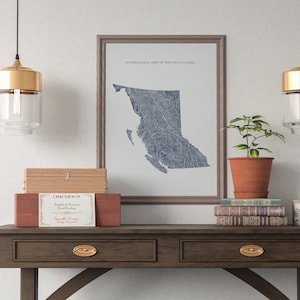 以下が含まれることがあります： 青と白の水文図のデザインが施された、ブリティッシュコロンビアの地図が額装されています。地図は、白い壁に、2つの金のペンダントライト、木製の箱、鉢植えの植物、本が積まれたテーブルの上にかけられています。