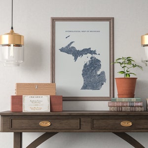 Op de afbeelding: Een ingelijste hydrologische kaart van Michigan in blauw- en grijstinten hangt boven een houten tafel. De tafel is versierd met boeken, een plant in een pot en decoratieve dozen. Twee hanglampen met gouden accenten en glazen kappen hangen boven de tafel.
