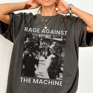 May include: Dark grey t-shirt with the text "RAGE AGAINST THE MACHINE" above and below a black and white image of people in a street. The shirt has short sleeves and a loose fit.