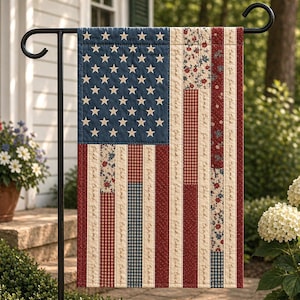 May include: A garden flag with a quilted design of the American flag. The flag features a blue field with white stars, red and white stripes, and floral and plaid accents. The flag is displayed on a black metal stand.