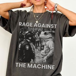 Könnte beinhalten: Dunkelgraues T-Shirt mit dem Text "RAGE AGAINST THE MACHINE" über einer Schwarz-Weiß-Grafik. Die Grafik zeigt einen Polizisten und einen Frosch mit Kapuzenpullover. Das T-Shirt hat eine übergroße Passform.