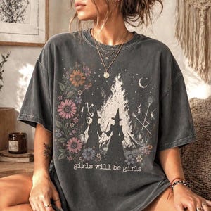 May include: Dark gray t-shirt featuring a floral and witch-themed graphic. The design includes two witch silhouettes, a bonfire, a crescent moon, and the text "girls will be girls". The shirt has a vintage, worn look.