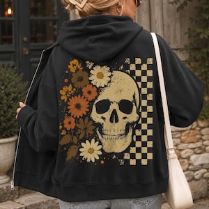 May include: A black zip-up hoodie featuring a large graphic on the back. The design displays a beige human skull partially surrounded by orange, yellow, and brown flowers on the left side and a beige and black checkered pattern on the right side.