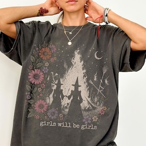 May include: Dark gray t-shirt with a graphic featuring two witch silhouettes, a bonfire, and floral accents. The text "girls will be girls" is printed below the graphic. The shirt has short sleeves and a relaxed fit.