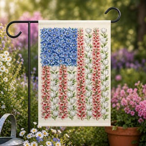 May include: A garden flag featuring a floral American flag design. The flag's blue field is composed of blue flowers, while the stripes are made of red and white flowers. The flag is displayed on a black metal stand in a garden setting.