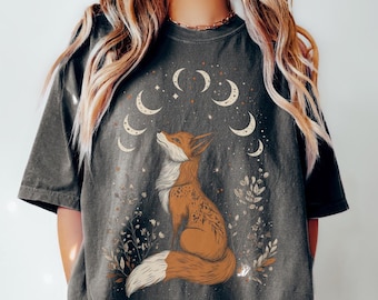May include: Dark gray t-shirt featuring a seated fox illustration with a crescent moon design. The fox is brown and white, surrounded by floral elements. The shirt has short sleeves and a relaxed fit.