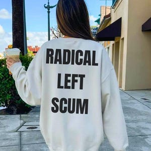 Peut inclure: Sweat-shirt couleur sable avec l'inscription "RADICAL LEFT SCUM" en lettres noires audacieuses dans le dos. Le mot "SAND" est imprimé en noir en bas à droite. Le sweat-shirt est de style décontracté.