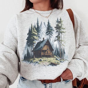 May include: A light gray sweatshirt featuring a cabin scene in a forest with tall green trees and a mountain backdrop. The cabin is brown with a dark blue roof. The sweatshirt has long sleeves and a crew neck.