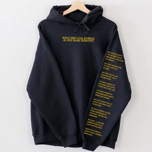May include: Black hoodie with the text "WHAT THEY CALL RADICAL IS JUST BASIC HUMANITY" in yellow on the front. The left sleeve has additional text in yellow. The hoodie is hanging on a wooden hanger.