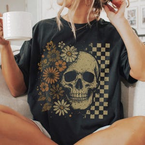May include: Black t-shirt featuring a skull design with floral accents and a checkered pattern. The skull is in the center, with flowers on the left side. The shirt is a dark color and has a vintage look.