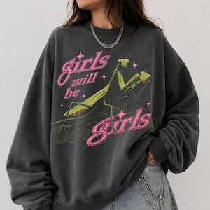 May include: A dark heather grey crewneck sweatshirt featuring a graphic print of a green praying mantis. The text "girls will be girls" is printed in a bright pink cursive font, accented by small pink star shapes across the chest area.