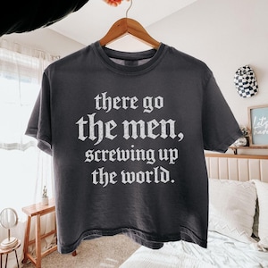 Puede incluir: Camiseta gris oscuro con el texto "there go the men, screwing up the world" en blanco, con fuente gótica. La camiseta está colgada en una percha de madera.