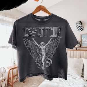 Peut inclure: T-shirt gris foncé avec le nom du groupe "Led Zeppelin" en blanc. Le t-shirt présente un dessin d'une femme ailée. Le t-shirt est suspendu à un cintre en bois.