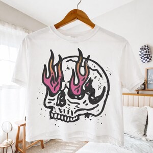 May include: White t-shirt featuring a black and pink skull graphic with flames emanating from the eye sockets. The design is a bold, graphic illustration. The shirt is hanging on a wooden hanger.