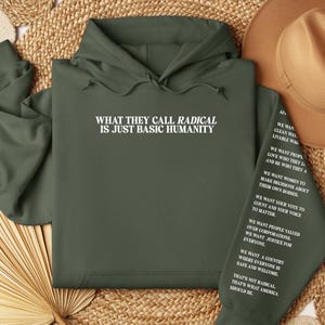 May include: An olive green hoodie with white text on the front that reads "WHAT THEY CALL RADICAL IS JUST BASIC HUMANITY." The left sleeve has additional white text. The hoodie is displayed on a woven surface.