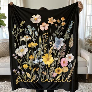 May include: Black blanket featuring a colorful floral design with yellow, white, pink, and blue flowers. The blanket has the words "Fuck Cancer" in a cursive font at the bottom. The blanket is held up by a person.